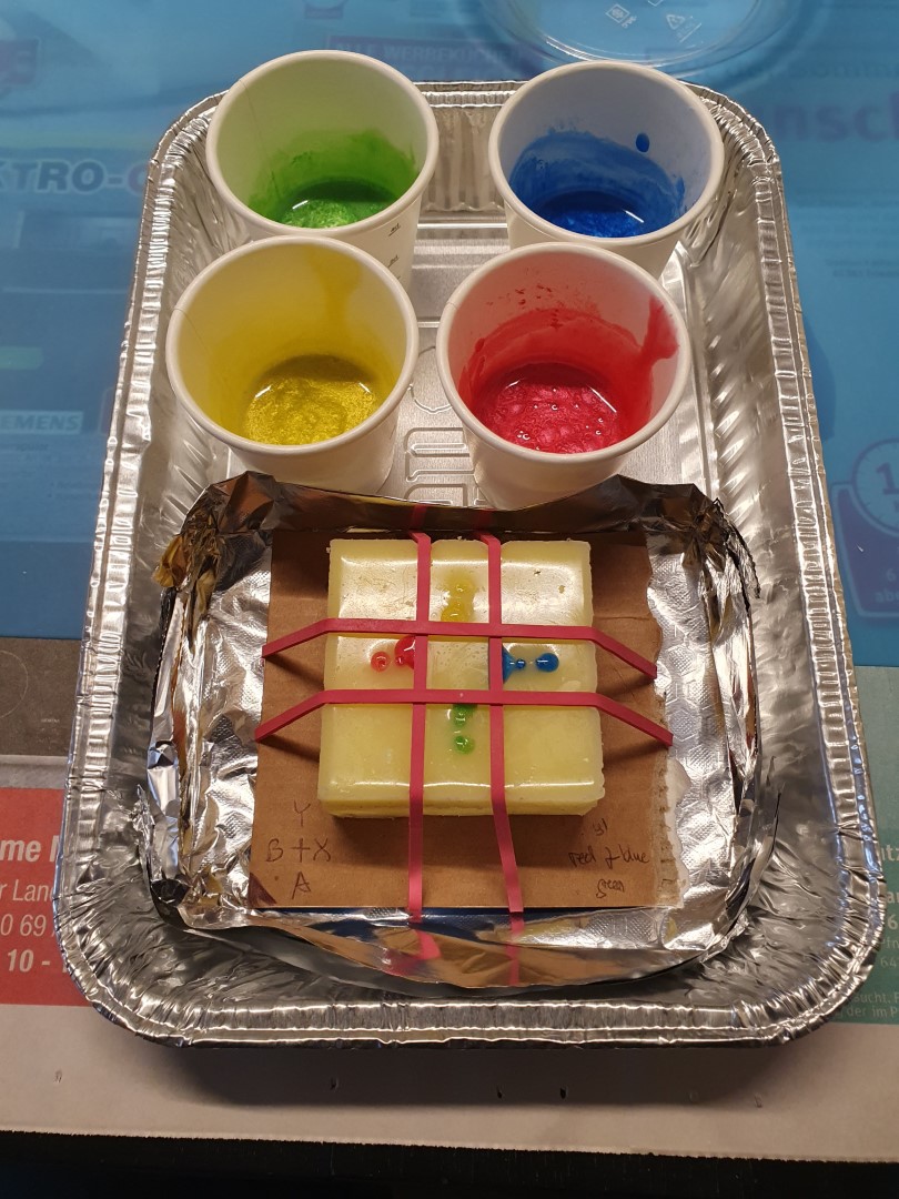 The button mold filled with the colored resin, the four small paper cups holding the unused resin sitting next to it in an aluminium tray.