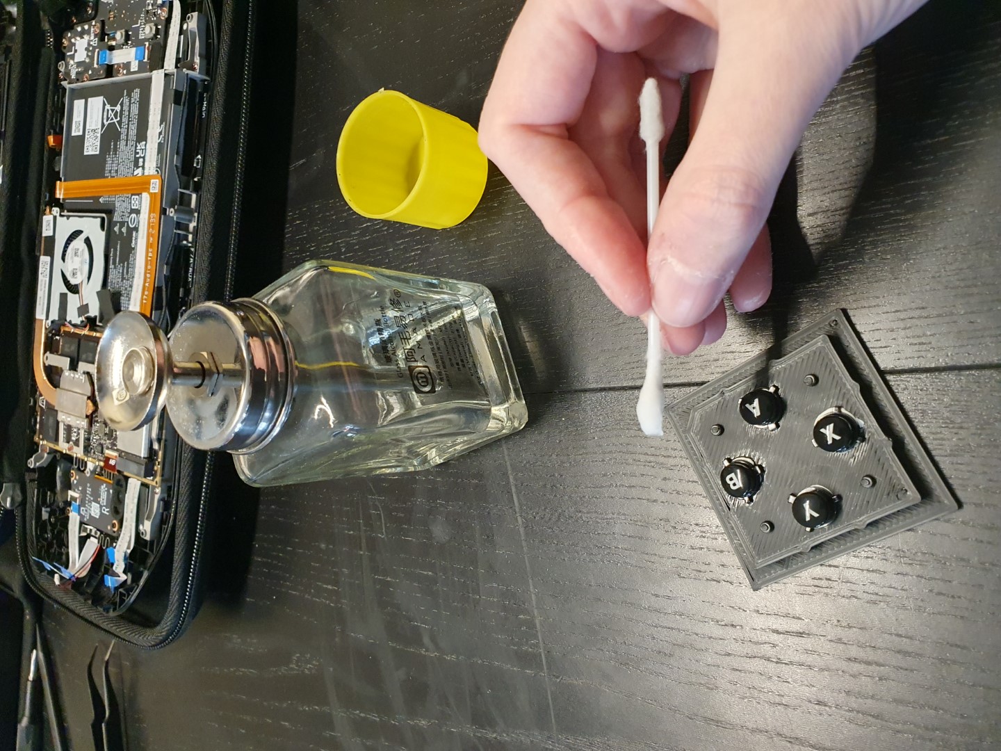 The four action buttons mounted to the keyed mold case bottom. My hand hovering over them with a q-tip, a bottle of isopropyl alcohol in reach. In the background the disassembled deck.