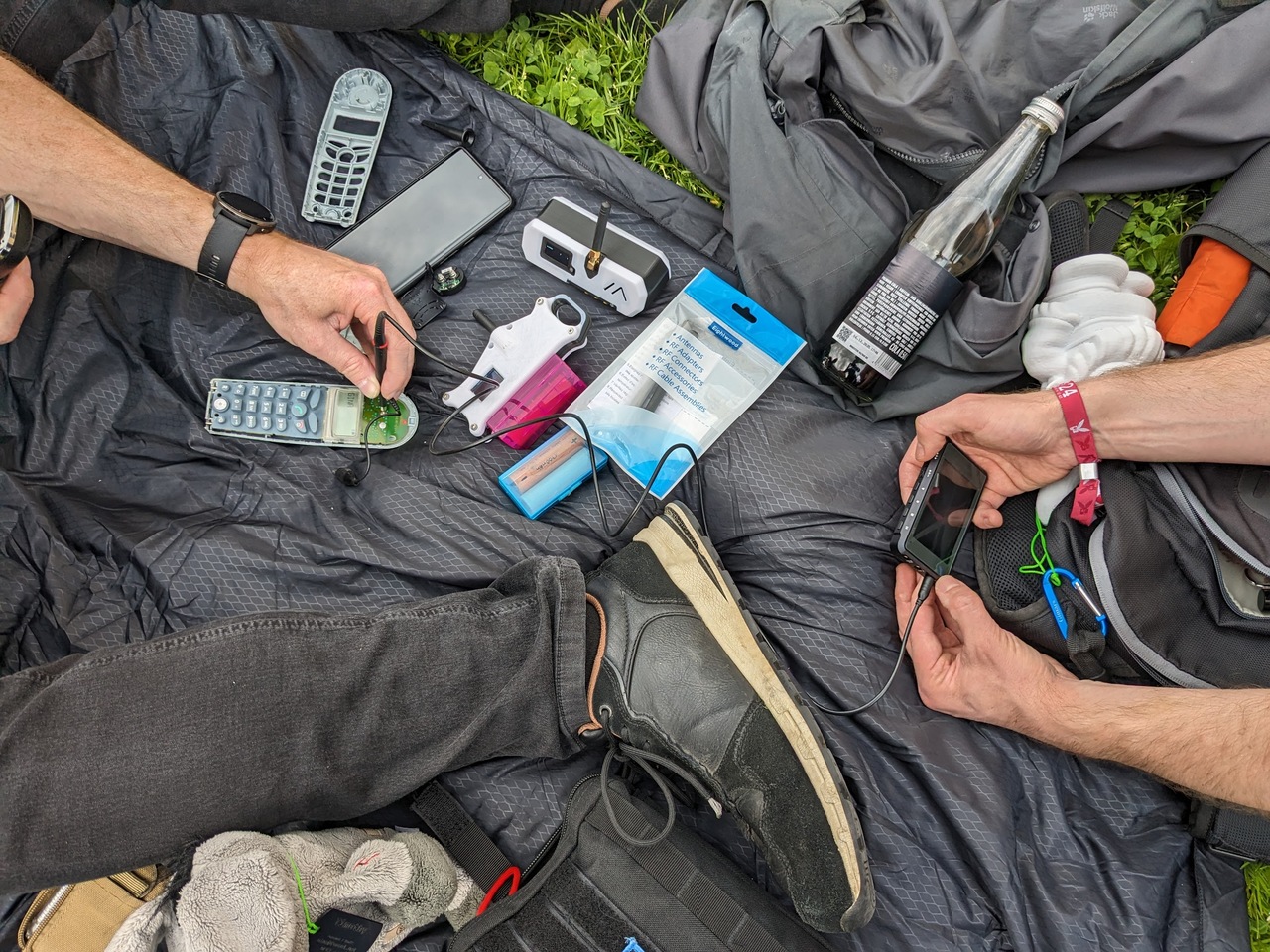 A picnic blanket with an opened up DECT phone, soldering tooling, a partial Datenzwerg, and some more electronics. Also - someone&rsquo;s leg and someone else&rsquo;s hands holding a portable oscilloscope